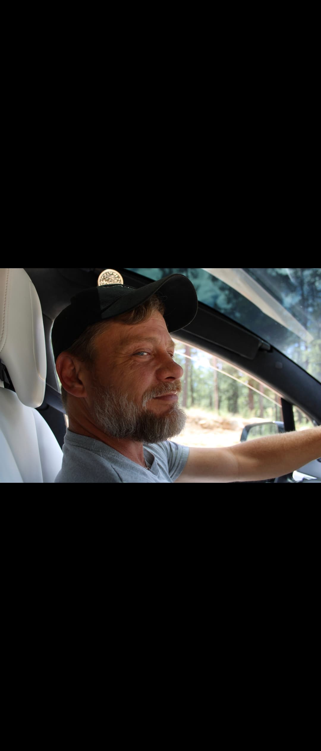 Bearded man in a black cap smiles from the driver's seat of a car.